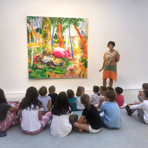 Vista frontal de María frente a un grupo de niños explicando un cuadro en una galería.